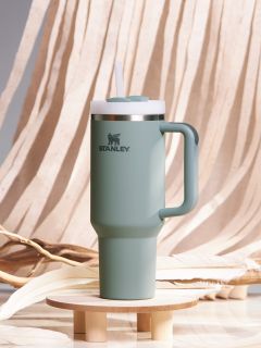 Un vaso de viaje Stanley verde con asa y tapa de pajita está sobre una pequeña base de madera. El fondo consiste en una tela beige que fluye y ramas decorativas.