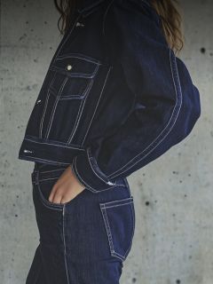 Una mujer con gorro blanco, camiseta blanca y vaqueros azules posa con el brazo extendido en una habitación moderna y blanca con flores encima.