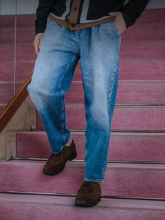 Un hombre con una camiseta blanca, una chaqueta vaquera oscura y vaqueros está de pie en una barandilla apoyada en una pared de hormigón y una valla, con un toldo amarillo encima.