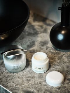 Dos tarros abiertos de crema facial, Sensai y La Mer, sobre una superficie de mármol gris junto a un jarrón negro y un lavabo.