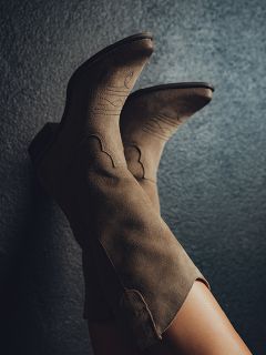 Primer plano de las piernas de una persona con botas de vaquero de ante color topo con costuras decorativas, apoyadas contra una pared texturizada de color gris oscuro.