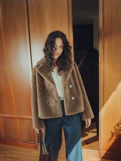 Una mujer con cabello rizado que lleva un abrigo de piel sintética marrón, una blusa blanca y vaqueros de pierna ancha está de pie en un interior con un bolso marrón en la mano, con paneles de madera y luz solar al fondo.