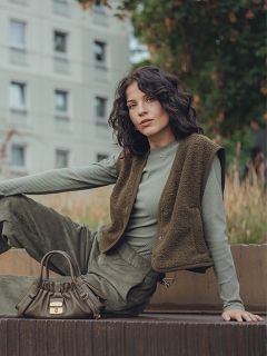 Una mujer con cabello oscuro ondulado está sentada al aire libre en una superficie elevada, vistiendo una blusa de canalé verde de manga larga, un chaleco color oliva y pantalones a juego. Tiene un pequeño bolso de mano color oliva a su lado y un edificio residencial al fondo.