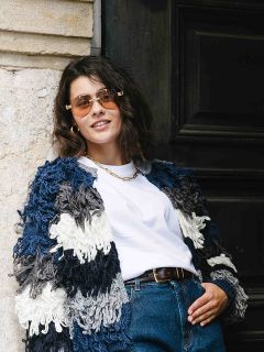 Una mujer con cabello castaño corto, que lleva gafas de sol, una chaqueta de punto gruesa azul y blanca, una camiseta blanca y vaqueros azules, está de pie casualmente frente a una pared de piedra y una puerta oscura, sonriendo con confianza.
