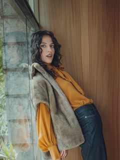 Una mujer con cabello oscuro ondulado está apoyada contra una pared de madera junto a una ventana. Lleva una blusa amarilla con mangas abullonadas, un chaleco de piel marrón y vaqueros oscuros. Su expresión es relajada mientras mira a la cámara.