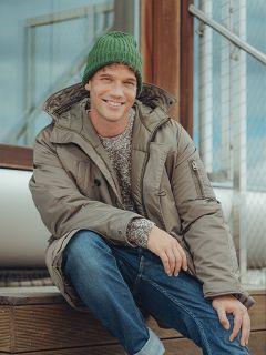 Un hombre sonriente, con un gorro de lana verde, un abrigo de invierno gris, vaqueros azules y un jersey estampado, está sentado en una escalera de madera al aire libre junto a una barandilla de metal.