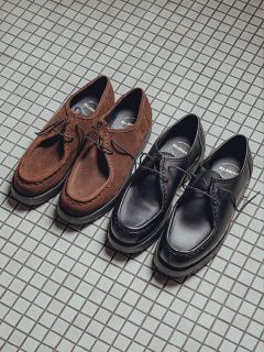 Dos pares de zapatos con cordones están uno al lado del otro sobre un suelo de baldosas: un par de ante marrón y el otro de cuero negro. Ambos zapatos tienen un diseño similar de estilo mocasín.
