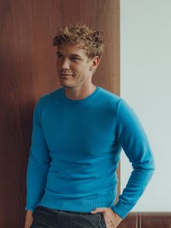 Un hombre con pelo rubio corto y rizado, que lleva un jersey azul claro y pantalones oscuros, está de pie en el interior junto a una pared de madera y blanca, y mira ligeramente hacia un lado con una expresión relajada.