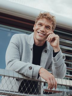 Un hombre sonriente con cabello rubio rizado se apoya casualmente en una valla de tela metálica, lleva un abrigo gris claro sobre una camisa negra y tiene un edificio moderno al fondo.