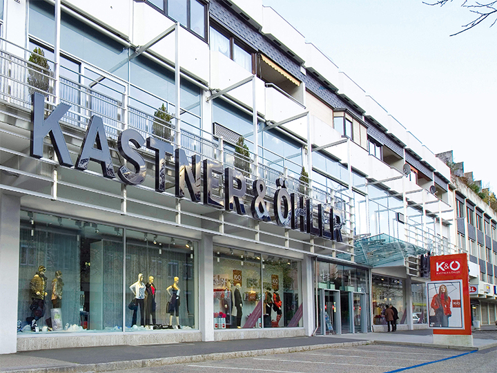 La imagen muestra la fachada de la tienda Kastner & Öhler con un gran logotipo rojo y blanco en el lado izquierdo y una moderna fachada de cristal. En las ventanas, debajo del letrero distintivo de la tienda, hay maniquíes con ropa elegante.