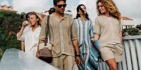 Un grupo de cinco personas se toma un selfie al aire libre cerca del agua, con árboles y una puesta de sol de fondo. Sonríen y posan juguetonamente, con ropa informal. Encima de ellos se ve el texto "KnowledgeCotton Apparel" y un logo de búho.