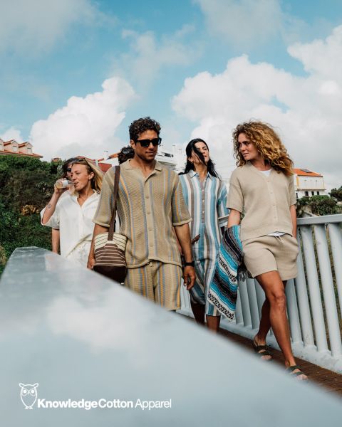 Un grupo de cinco personas se toma un selfie al aire libre cerca del agua, con árboles y una puesta de sol de fondo. Sonríen y posan juguetonamente, con ropa informal. Encima de ellos se ve el texto "KnowledgeCotton Apparel" y un logo de búho.