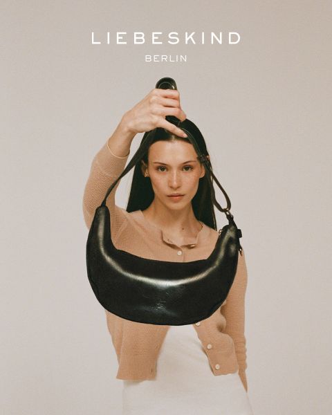 Una mujer sonriente con una camisa negra de manga larga y jeans lleva un bolso marrón claro sobre el hombro. El texto "Liebeskind Berlin" aparece en la parte inferior de la imagen.