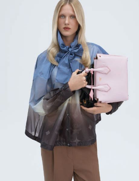 Un bolso de diseño verde con un sutil patrón de monograma descansa sobre una superficie gris, con un objeto geométrico azul en el primer plano inferior y una sombra proyectada en el fondo.