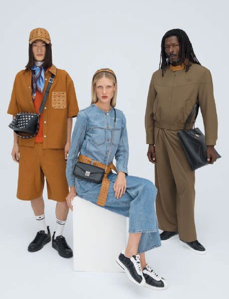 Un bolso de diseño MCM azul con un patrón de logotipo blanco está sobre escalones de madera adornados con coloridos llaveros de animales y objetos, incluyendo un cohete, un conejo y un oso.