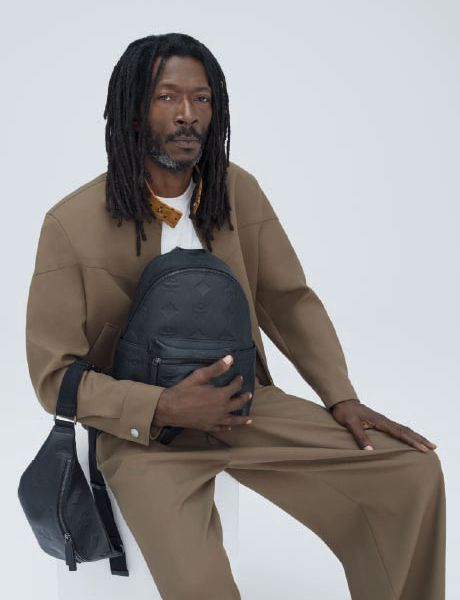 Una mochila de diseño verde y un bolso bandolera a juego están sobre una pila de cajas naranjas y marrones frente a una pared con textura beige y un suelo de baldosas.