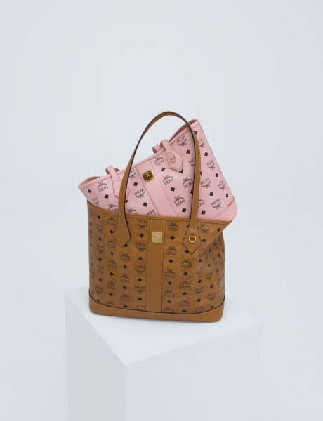 Una mujer con una chaqueta de shearling marrón claro, pantalones cortos negros y botas está de pie en un escalón de piedra y sostiene un bolso grande. En el fondo, un hombre con ropa colorida parece flotar horizontalmente sobre los escalones. Detrás de ellos se ven árboles.