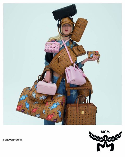 Tres mujeres con abrigos de color marrón claro y sombreros naranjas cruzan una calle, llevando bolsos azules de MCM con un logo blanco de MCM en primer plano. La imagen tiene un efecto espejo que hace que la escena parezca duplicada.