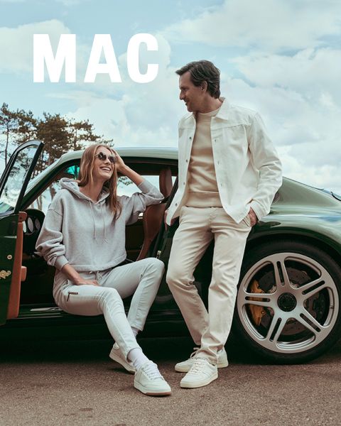 Una mujer con una chaqueta de piel y pantalones anchos está de pie junto a un hombre con una chaqueta beige y pantalones caqui. Ambos posan con confianza al aire libre frente a una barandilla de piedra bajo un cielo azul. La palabra "MAC" aparece en la parte inferior.