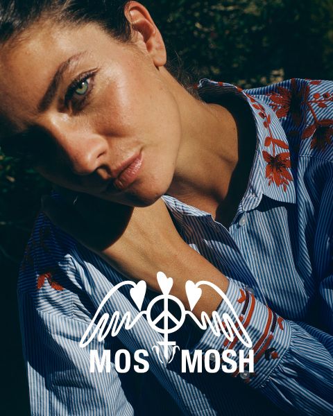 Una mujer con cabello rubio largo y ondulado lleva una camisa verde con patrones blancos. Mira por encima de su hombro y está de pie frente a un edificio moderno. El logo de "MOS MOSH" se encuentra en la esquina superior izquierda.