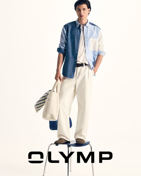 Un hombre con pantalones de traje, camisa a cuadros y corbata con un jersey drapeado sobre los hombros está de pie frente a un fondo azul oscuro, junto al cual se puede leer la palabra "OLYMP".