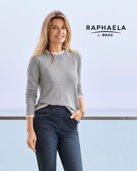 Una mujer con cabello largo y rubio, que lleva una camisa gris de manga larga y vaqueros oscuros, está de pie sonriendo junto a una barandilla con un fondo azul claro. Junto a ella aparece el texto "EUREX de BRAX".