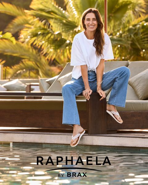 Una mujer con cabello largo y rubio, que lleva una camisa gris de manga larga y vaqueros oscuros, está de pie sonriendo junto a una barandilla con un fondo azul claro. Junto a ella aparece el texto "EUREX de BRAX".