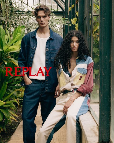 Un hombre y una mujer, vestidos con ropa oscura y elegante, están sentados en una escalera de madera en un interior. La palabra "REPLAY" aparece prominentemente en el centro de la imagen en letras mayúsculas rojas.