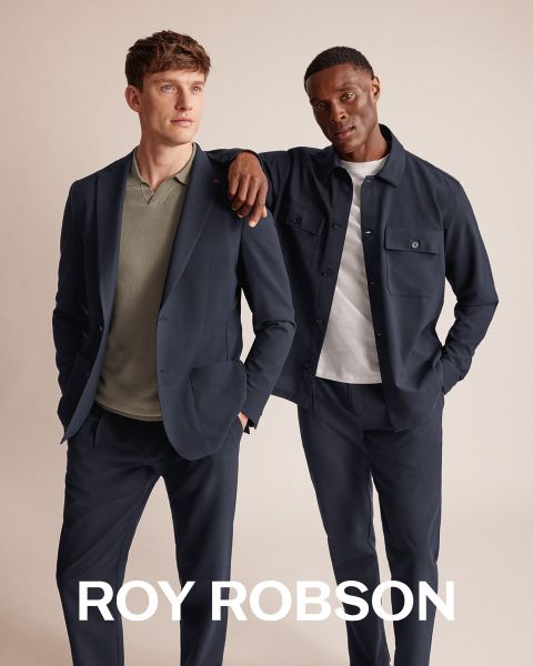 Un hombre está de pie al aire libre frente a montañas nevadas y lleva un abrigo de pata de gallo gris, un jersey gris y pantalones grises. El texto "ROY ROBSON" aparece en la parte inferior de la imagen.