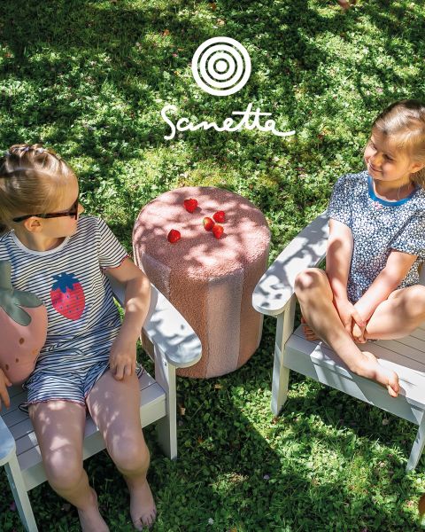 Cuatro niños sonrientes están de pie en un prado sosteniendo varas entre las que se extienden coloridas tiras de tela. Dos alpacas están cerca, y en el fondo se ven un cielo nublado y árboles. Arriba está escrito "Sanetta".