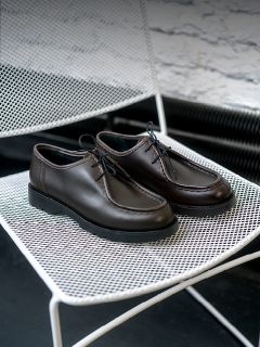 Persona con vaqueros remangados de pie sobre suelo arenoso cerca de cactus, con zapatillas blancas con detalles en beige y marrón y un logotipo en "V" en el lateral.