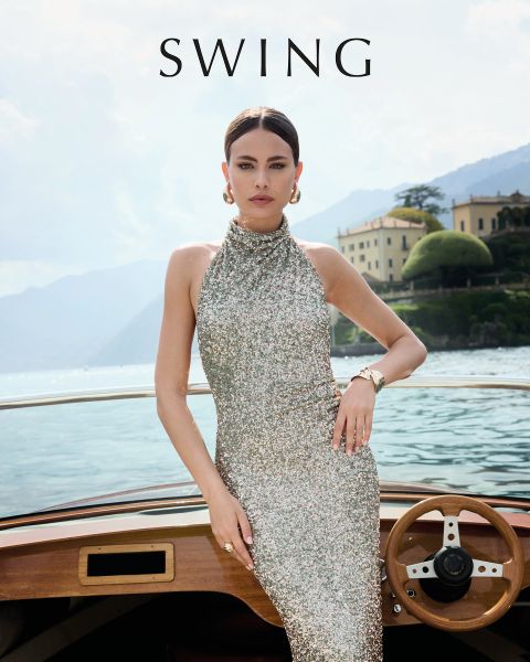 Una mujer con un vestido de seda verde salvia está sentada en una silla color crema frente a un fondo floral. Lleva tacones dorados, pendientes llamativos y una pulsera. La palabra "SWING" aparece en la parte inferior de la imagen.