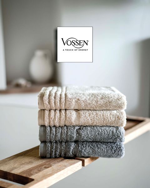 Una mujer con cabello rubio corto, envuelta en una toalla gris, está sentada en un sofá claro y mira a la cámara. En la esquina inferior izquierda se ve el logo de Vossen.
