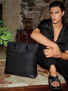 Una persona de cabello oscuro está sentada en una superficie de mármol, vestida con una camisa oscura abotonada y pantalones. Está descalza, con un pie sobre el otro, y lleva sandalias negras. A su lado hay una gran bolsa de cuero negra. En el fondo se ven una pared y muebles de estilo vintage.