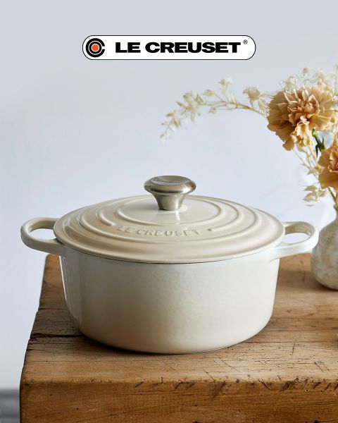 Una escena de cocina con una olla de cocción lenta roja, una sartén antiadherente, un juego de ollas apilables de acero inoxidable, una olla negra y un cuenco de tomates cherry sobre una encimera de madera clara frente a un protector contra salpicaduras de azulejos blancos. El logotipo de Le Creuset es visible.