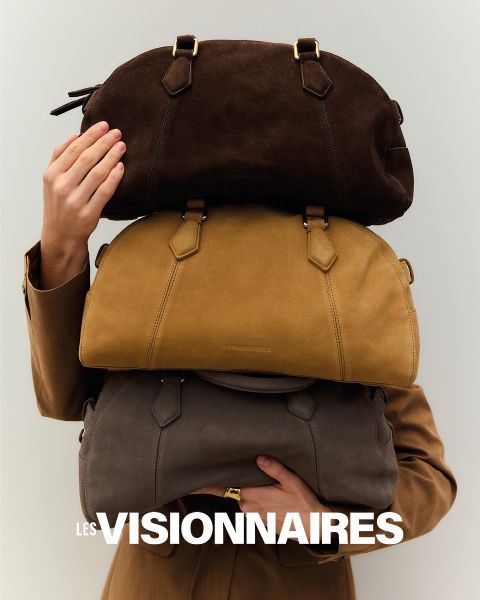 Una persona con un abrigo marrón levanta una mano para cubrir parcialmente su rostro. Está de pie frente a un fondo oscuro. En la parte superior de la imagen, las palabras "Les Visionnaires" están escritas en letras blancas y negritas.