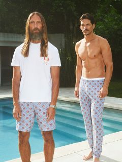 Un hombre sin camisa con un blazer negro y pantalones cortos oscuros está sentado en una silla blanca en unos acantilados rocosos junto al mar, sosteniendo una copa de vino blanco en la mano, mientras las olas rompen en el fondo.