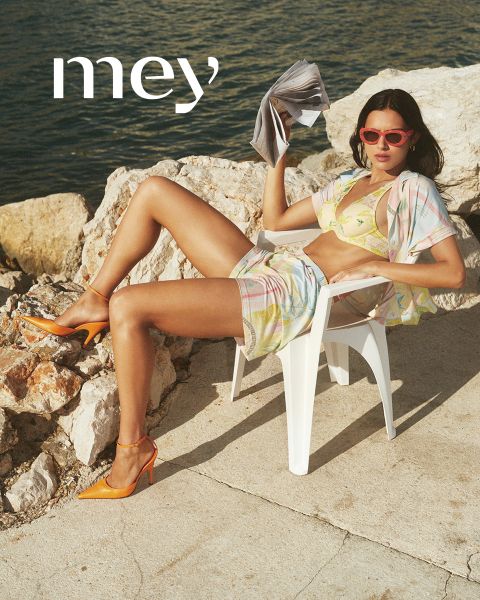 Un hombre en calzoncillos bóxer negros y una mujer en una camiseta sin mangas blanca y ropa interior están de pie uno al lado del otro al aire libre junto a una barandilla, con el mar y el cielo nublado de fondo. La palabra "mey" está en la parte inferior.