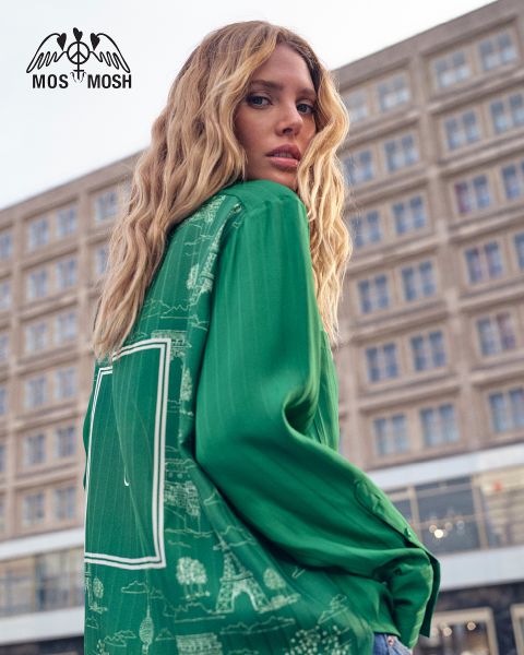 Una mujer con cabello rubio largo y ondulado lleva una camisa verde con patrones blancos. Mira por encima de su hombro y está de pie frente a un edificio moderno. El logo de "MOS MOSH" se encuentra en la esquina superior izquierda.