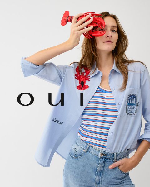 Una mujer que lleva un jersey rojo con un corazón blanco, vaqueros azules y un gorro blanco posa delante de una pared clara. En la parte inferior de la imagen está la palabra "oui" en letras blancas.