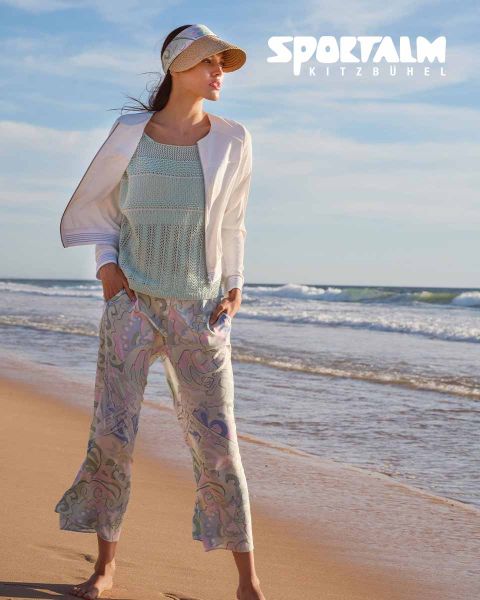 Una mujer está de pie en una playa. Lleva un top de punto azul claro, una chaqueta blanca y unos pantalones estampados. Lleva una visera y mira hacia el mar. En la esquina superior derecha pone "SPORTALM KITZBÜHEL".