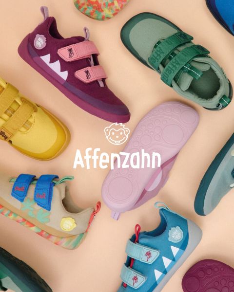 Un niño con un suéter naranja brillante y pantalones a rayas está sentado en una superficie colorida y señala un avión de papel estilo cómic en el cielo. Sonríe y está rodeado de nubes esponjosas. En la esquina pone "Affenzahn".