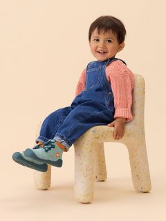 Un niño pequeño con camisa a rayas y mono de mezclilla está sentado con una pierna al aire libre, mostrando la suela azul texturizada de su zapato.