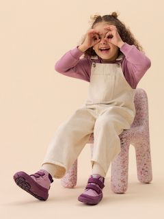 Un niño sonriente con un gorro verde, camisa a rayas y chaleco está sentado sobre un bloque de color melocotón frente a un fondo amarillo, vistiendo botas y pantalones claros.