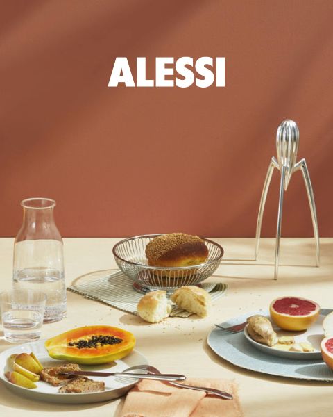 Dos hueveras geométricas modernas con huevos están sobre una bandeja reflectante, junto con un cuenco de vidrio y una cuchara. En la parte inferior central de la imagen se lee "ALESSI" en negrita.