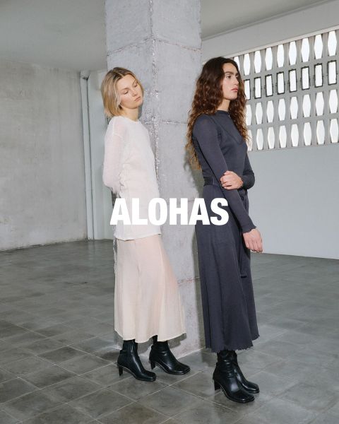 Una mujer con un top blanco que deja ver el abdomen y pantalones beige con detalles naranjas está sentada en un taburete. Se encuentra en una habitación rústica con grandes ventanas. En el centro se ve el texto "ALOHAS".