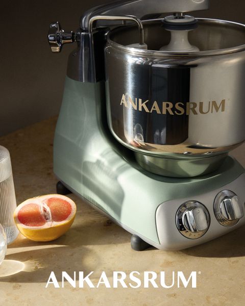 Una brillante batidora Ankarsrum con un cuenco de acero inoxidable y una base rosa se presenta sobre una superficie rosa a juego, con el nombre de la marca "ANKARSRUM" visible tanto en el cuenco como en el fondo.