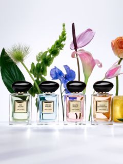 Cuatro frascos de perfume se exhiben sobre y alrededor de un pedestal beige. Los frascos son cuadrados con tapas redondeadas y en colores negro y rojo. Una pared blanca ondulada y texturizada sirve de fondo.