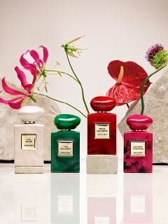 Ocho frascos de perfume en varios colores, incluyendo rojo, verde, azul, rosa y negro, están dispuestos sobre pedestales beige frente a un fondo claro. Cada frasco tiene una etiqueta con letras doradas.