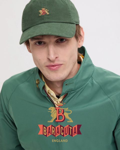 Una persona con cabello corto y oscuro y un pendiente, que lleva una chaqueta oscura sobre una camisa blanca, mira a la cámara. El interior de la chaqueta es de cuadros rojos. En la parte delantera se muestra el logotipo de Baracuta con un león y una "B".